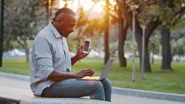 Afroameričan zkušený profesionál mluví po telefonu pomocí notebooku, sedí venku. Starší podnikatel podnikatel učinit mobilní hovor potvrzující on-line doručení objednávky webové stránky — Stock video