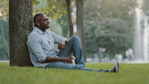 Feliz anciano africano negro hombre de negocios con auriculares inalámbricos sentado en la hierba en el parque de la ciudad escuchar música en el teléfono inteligente, hombre alegre de la edad de jubilación en los auriculares modernos disfrutando del descanso al aire libre — Vídeo de stock
