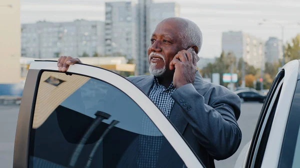 Olgun Afro-Amerikalı iş adamı dışarıda arabanın yanında duruyor. Telefonlara cevap veriyor. İş görüşmeleri yapıyor. — Stok fotoğraf