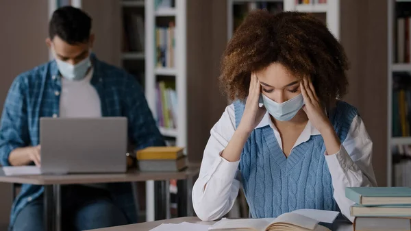 Dois alunos em máscara protetora sentados em sala de aula na biblioteca da universidade fazendo lição de casa preparação exame jovem à procura de informações em livro segurando cabeça mãos cara dor de cabeça estudando no laptop — Fotografia de Stock