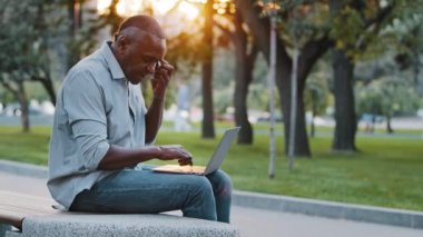 Afrikalı Amerikalı bir adam müşteriyle uzaktan telefon görüşmeleri yaparak profesyonel yardım alma tecrübesi edindi. Etnik yaşlı iş adamı dizüstü bilgisayar kullanıyor, şehir parkında oturuyor.