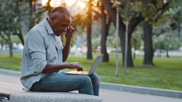 Homem afro-americano experiente profissional segurando smartphones fala com o cliente distantemente fornecendo ajuda por telefone. Homem de negócios idoso étnico usando computador portátil, sentado ao ar livre no parque da cidade — Vídeo de Stock