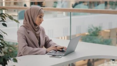 Odaklanmış ciddi Arap iş kadını profesyonel girişimci ofis masasında oturmuş dizüstü bilgisayara bakıyor online kurs müslüman kız serbest çalışan bilgisayar aygıtı kullanıyor.