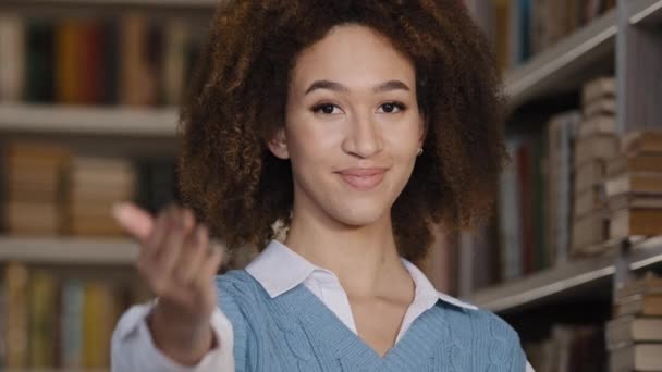 Close-up jong Afrikaans amerikaans vrouw student staan in de openbare universiteit bibliotheek kijken naar camera glimlachen maakt gebaar uitnodiging uitnodigen om goed college onderwijs nodigt uit om boeken te lezen — Stockvideo