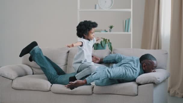 Adult african american father lying stomach on sofa little daughter plays sits on dads back kid girl pretends to fly on plane sitting on daddy spread arms to side mature man fun playing with child — Stock Video