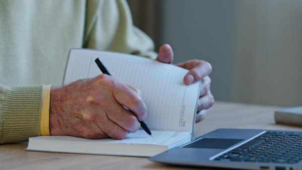 Close-up hånd ældre kaukasiske mand skrive noter i personlige papir planner på skrivebordet uigenkendelige alderen mandlige gør i notesbog to-do liste daglige tidsplan skrive oplysninger fra internettet i laptop – Stock-video
