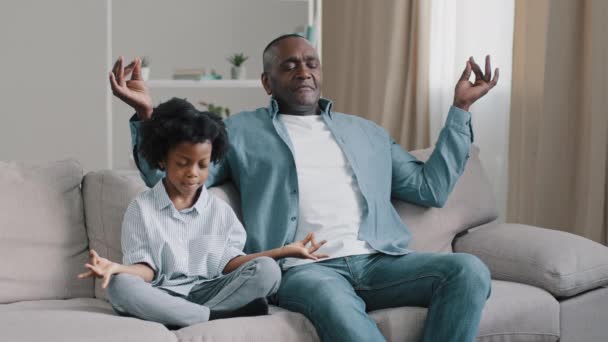 Afrikalı Amerikalı baba ve çocuk kanepede gözleri kapalı oturuyor. Olgun baba küçük kızına meditasyon yapmayı öğretiyor. Duygularını kontrol etmeyi. — Stok video
