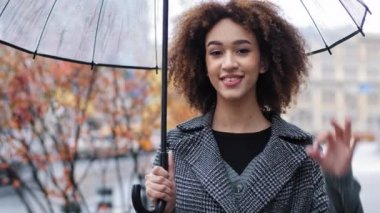 Güzel, mutlu, mutlu, kıvırcık, Afro-Amerikan kadın, şeffaf şemsiyeli, yağmurlu sonbahar şehrinde kameraya bakıp gülümsüyor.