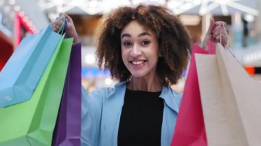 Yakın plan, mutlu Afro-Amerikan kadın elinde parlak alışveriş çantaları paketler indirimlerden zevk alıyor indirimli kameralara bakıp bayram indirimi için alınmış hediyeleri geniş bir gülümsemeyle gösteriyor.