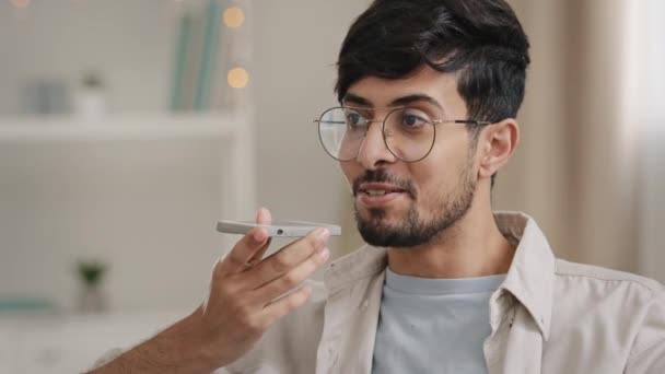 Amistoso joven árabe grabando mensaje de reconocimiento de voz en altavoz sentado en casa. Chico barbudo con gafas usando asistente virtual conjuntos de aplicaciones recordatorio en el teléfono móvil en interiores dicta palabras — Vídeo de stock