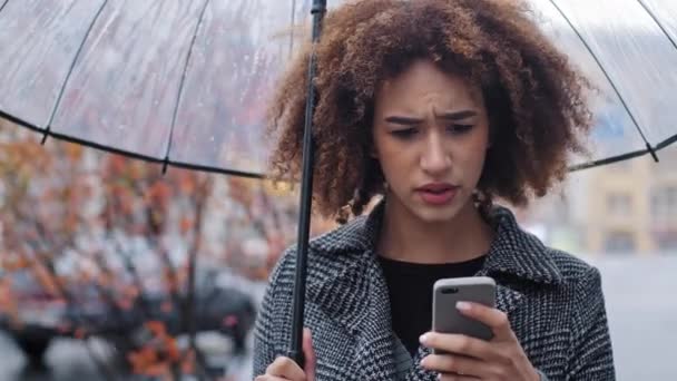 雨の中で秋の通りに立つ透明な傘を持つアフリカ系アメリカ人の女性は、インターネットの悪いニュースと低バッテリーの問題を心配して携帯電話の動揺悲しみを見て拒否されます — ストック動画