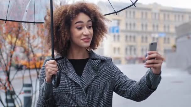 Amichevole sorridente ragazza riccia donna afro-americana blogger vlogger turista in possesso di ombrello trasparente in piedi in città in autunno pioggia registrazione video blog vlog online conferenza chiamata a distanza — Video Stock