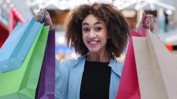 Close-up zadowolony szczęśliwy Afryki amerykanka dziewczyna gospodarstwa jasne zakupy torby zakupy pakiety cieszyć się zniżki patrząc na aparat uśmiechając się szeroko pokazując zakupione prezenty na wakacje koncepcji sprzedaży — Wideo stockowe