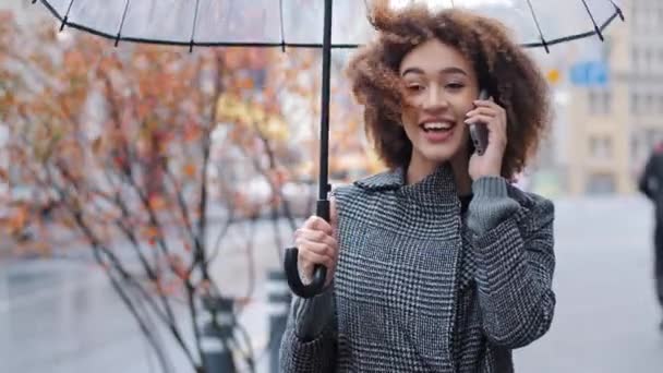 Ευτυχισμένο ενεργό φιλικό σγουρά μαλλιά κορίτσι της Αφρικής αμερικανική γυναίκα με διαφανή ομπρέλα στην πόλη σε θυελλώδη βροχερό καιρό απαντώντας κλήση μιλάμε στο κινητό τηλέφωνο καλώντας ταξί συνομιλία με smartphone — Αρχείο Βίντεο