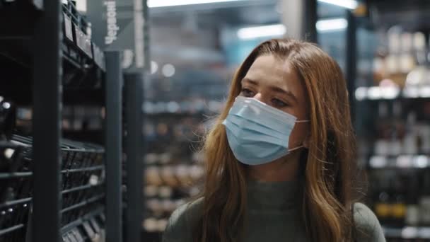 Close-up joven mujer caucásica chica consumidor comprador lleva máscara médica durante la pandemia coronavirus caminar a través de la tienda elegir botella de vino mira en los estantes con alcohol, compras en cuarentena — Vídeo de stock