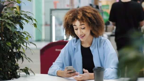 Junge schöne Mädchen Teenager Millennial Afroamerikanerin mit lockigem Haar sitzt im Café und schaut auf Handy freut sich gewinnt bekommt Sieg gutes Ergebnis gewinnt in Online-Spiel Glück Erfolg Triumph — Stockvideo