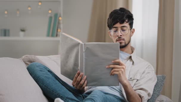 Árabe joven inteligente estudiante barbudo hombre en gafas se encuentra en el sofá en casa libro de lectura libro de texto aprender nuevo idioma estudiando la preparación para el examen de descanso lee disfrutando de ocio hobby en el apartamento — Vídeos de Stock