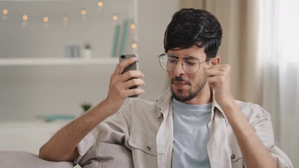 Joven 30s árabe barbudo hombre millennial chico en gafas se sienta en casa en sofá mirando el teléfono móvil haciendo sí gesto conseguir oferta ganar buenas noticias ganar victoria anuncio sentir feliz éxito — Vídeo de stock