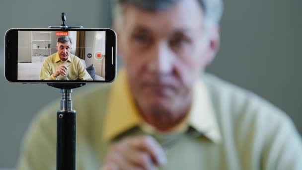 Close-up zaměření na smartphone starší muž provádí on-line vysílání záznam video blog na mobilní telefon sdílení myslel vážný starý profesor učit vzdáleně sedí v obývacím pokoji virtuální událost koncept — Stock video