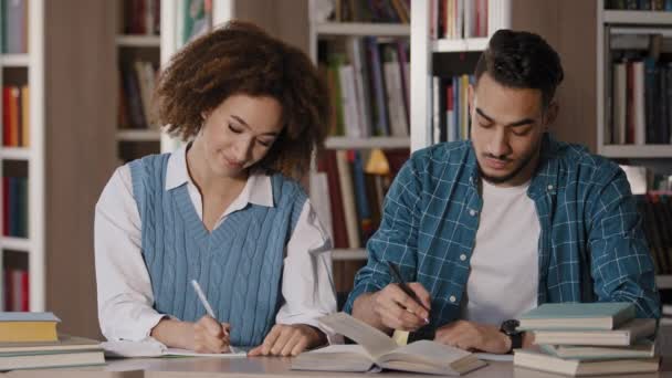 Dwoje uczniów młody chłopak i dziewczyna odrabiają lekcje siedząc przy biurku w bibliotece i pisząc notatki uważnie słuchając wykładowcy na lekcjach w klasie szkolnej koledzy z klasy rozmawiający uśmiechnięty — Wideo stockowe