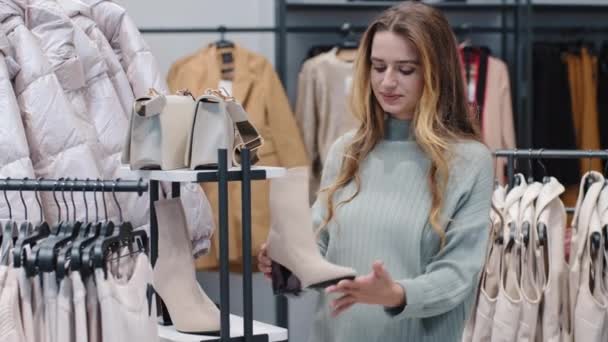 Stijlvol meisje vrouwelijke Kaukasische millennial shopper dame consument in kledingwinkel mode boetiek op zoek naar prijzen kiezen product houden knuffelen witte laars denken nadenken, winkelen concept — Stockvideo