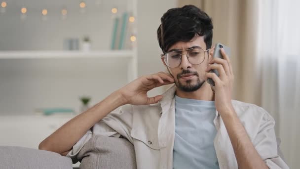 Junger arabischer bärtiger Mann Millennial Kerl mit Brille sitzt auf der heimischen Couch und spricht auf dem Handy Anrufbeantworter Sorgen über schlechte Netzwerkprobleme Mobilfunk-Problem kaputtes Smartphone-Mikrofon — Stockvideo