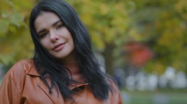 Güzel, zarif, esmer bir kadın sonbahar parkında duruyor, kilolu kız model açık havada kameraya bakıyor, utanarak aşağı bakıyor, gülümsüyor, çenesinin altından boynunu tutuyor.