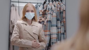 Koruyucu tıbbi maskeli genç bir kadın mağazada yeni kıyafetler seçiyor. Sonbahar ceketi giyip aynadaki beyaz kıza bakıyor. Pandemik karantina altındaki hafta sonu alışveriş konsepti sırasında kıyafet alıyor.
