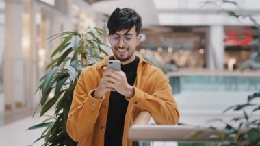 Genç ispanyol adam elinde telefonuyla duruyor gülümsüyor sosyal ağ üzerinden e-posta gönderiyor internet sayfası kontrolleri haberlerde akıllı telefondaki uygulamaları kullanıyor telefon üzerinden online mağazada sipariş veriyor