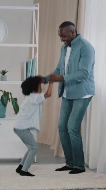 Dikey video Afro-Amerikan babası, küçük kızıyla eğleniyor dans ediyor, el ele tutuşuyor, gülüyor, sevimli kız zıplıyor, babasına yardım ediyor mutlu aile evde aktif olarak oynuyor.