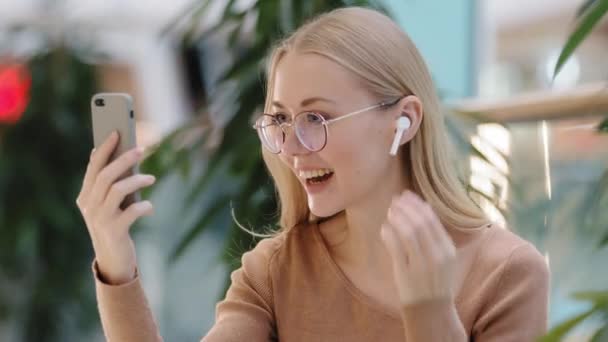 Emocionado joven atractiva chica milenaria con auriculares inalámbricos chat por aplicación de uso de videollamada en el teléfono mujer caucásica con gafas sonriendo felizmente reunión virtual comunicación remota tecnología moderna — Vídeos de Stock