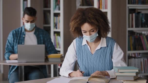 Két diák védő maszk ül osztályteremben egyetemi könyvtár csinál házi feladat vizsga előkészítése fiatal lány keres információt könyv fogása fej kéz fejfájás srác tanul laptop — Stock videók