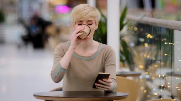 Middle aged adult woman caucasian lady with short haircut sitting at cafe table drinking coffee tea looking in mobile phone chatting browsing with smartphone buying online using internet app message — Stock Video