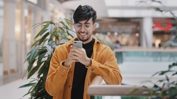 Junge hispanische Kerl steht in Mall mit Telefon lächelnd E-Mail senden auf sozialem Netzwerk surfen Web-Seite überprüft Nachrichten verwendet mobile Anwendung auf Smartphone Bestellung im Online-Shop am Telefon — Stockvideo