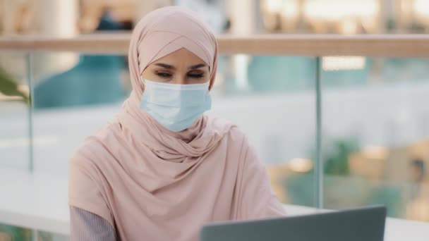 Joven mujer de negocios árabe en el hijab trabaja en el ordenador portátil mira alrededor chica musulmana baja máscara médica exhala con alivio arañazos divertidos nariz pone respirador en las caras mujer cansada de restricciones de cuarentena — Vídeos de Stock