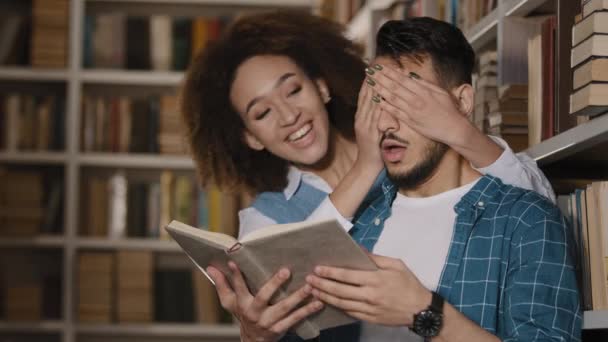Молодой испаноязычный студент-мужчина стоит в библиотеке, читает книгу, африканская американская девушка неожиданно подходит парень закрывает глаза удивляет друзей обнимает улыбается говорить общаться счастливы видеть друг друга — стоковое видео