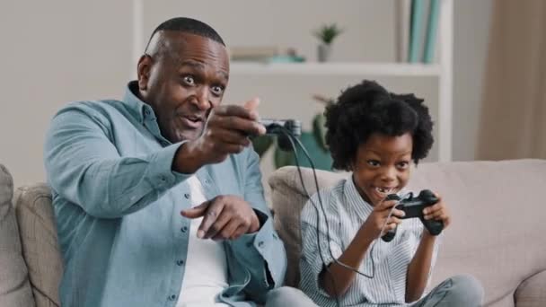 작은 딸을 데리고 콘솔 게임을 하는 미국 아프리카 계 미국인 아버지 조이스틱 컨트롤러 소파에 앉아 게임을 즐기며 웃는 아빠 — 비디오
