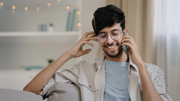 Бородатый мужчина в очках, сидящий дома на диване и разговаривающий по мобильному телефону, болтая с друзьями — стоковое фото