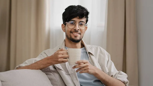 Arab szakállas Y-generációs férfi latin fickó szemüvegben otthon ül élvezi finom forró gyümölcs gyógynövény tea kávé ital kakaó ivás cappuccino ital azt mondja, igen pozitívan bólint fej hangulat koncepció — Stock Fotó