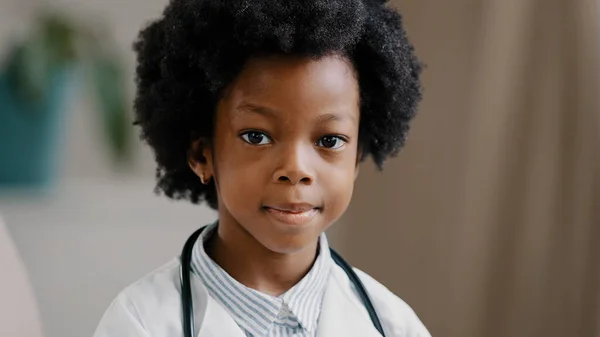 Cute serious african american kid girl in medical clothes dressed in white coat standing indoors posing looking at camera playing pretending to be doctor future profession concept close-up portrait — Stock Photo, Image