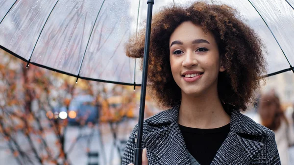 Afrikaans amerikaans meisje mooi twintiger jaren dame met krullend haar vrouwelijk model staan op straat stad herfst regen met transparante paraplu glimlachen tandheelkundige kijkend in afstand genieten van regenachtig weer — Stockfoto