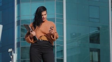 Genç İspanyol iş kadını arka planda duruyor. Telefon ekranına bakıyor. Akıllı telefondan e-posta gönderiyor.
