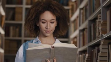 Genç Afrikalı Amerikalı kız öğrenci, devlet üniversitesi kütüphanesinde durmuş ilginç kitap hikayeleri okumakla ilgileniyor ders kitabına bakıyor edebiyat okumaktan keyif alıyor.