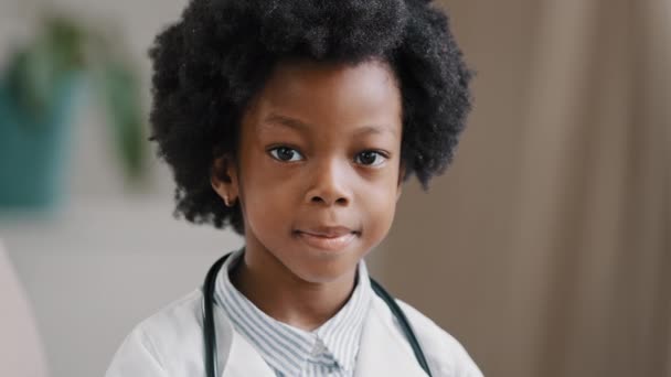 Aranyos komoly afro-amerikai gyerek lány orvosi ruhába öltözött fehér kabát áll beltérben pózol kamera játszik színlelt orvos jövőbeni szakma koncepció közelkép portré — Stock videók