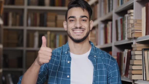 Крупный план счастливый молодой латиноамериканец студент, стоящий в публичной университетской библиотеке улыбаясь показывая большой палец вверх жест одобрения давая положительную обратную связь наслаждаться учебой в высшем учебном заведении успешное обучение — стоковое видео