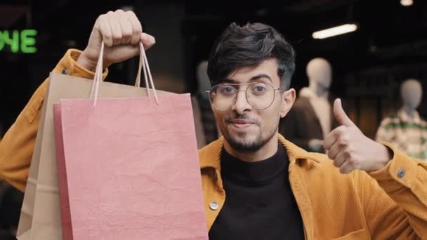 Yakın plan mutlu genç Arap adam giyim mağazasında gülümsüyor başparmağıyla jest onayı veriyor alışveriş için hediye çantalarını işaret ediyor indirimli satışla satın alınan yeni şanslı alışverişin keyfini çıkarıyor. — Stok video