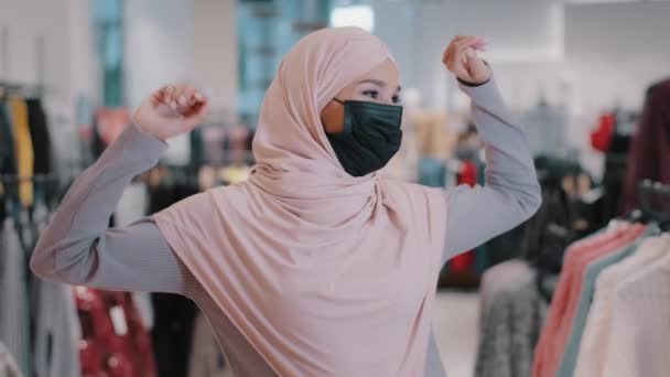 Feliz despreocupado ativo engraçado jovem árabe mulher comprador em hijab vestindo máscara médica protetora se movendo dinamicamente dança alegre se move para a música na loja de roupas se alegra na venda goza de desconto — Vídeo de Stock