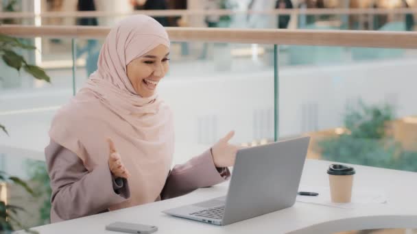 Feliz chica árabe joven en hijab sentado en la mesa respondiendo a una videollamada en el portátil saludando alegremente con la mano charlando con un amigo en la webcam hablando utilizando la aplicación informática se comunica de forma remota — Vídeo de stock