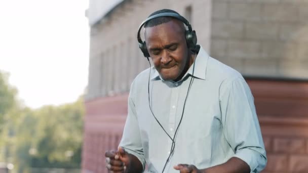 Nadšený šťastný starší muž nosí bezdrátové sluchátka baví poslouchat hudbu v smartphonu, tanec, veselá osoba důchodového věku v moderních sluchátkách předstírat hrát bicí venku — Stock video