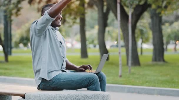 Adulto maduro afro-americano homem profissional usando computador portátil, sentado ao ar livre no parque da cidade. Etnia idoso empresário cumprimentando amigo acenando Olá ou adeus muito ocupado promete chamar — Vídeo de Stock